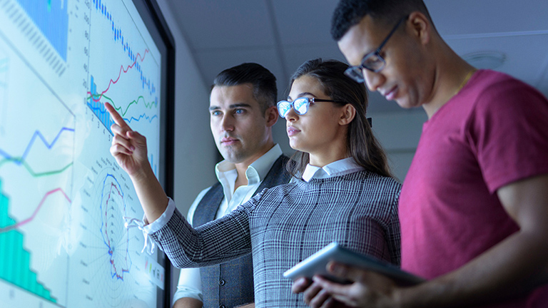 Three coworkers looking at various charts on a large wall screen