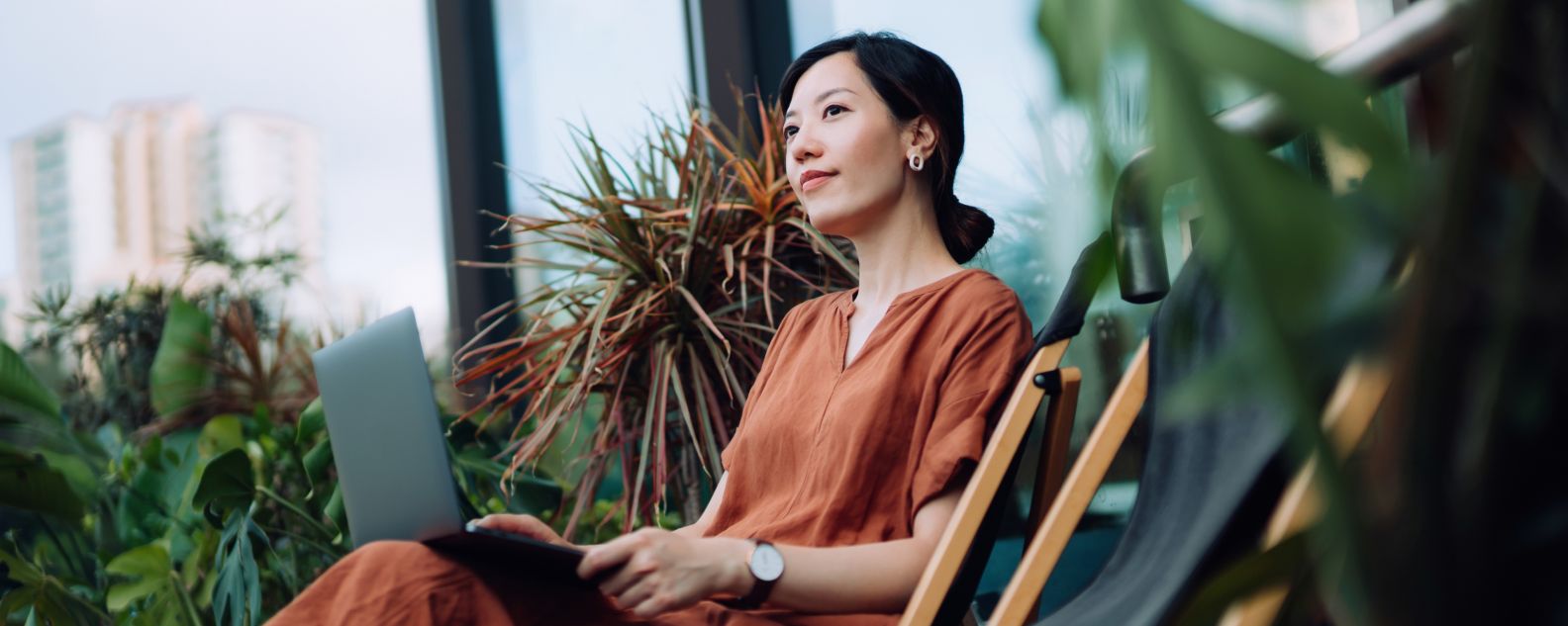 Contemplated young Asian woman looking away in thought while relaxing on deck chair using laptop in the backyard, surrounded by beautiful houseplants. Lifestyle and technology