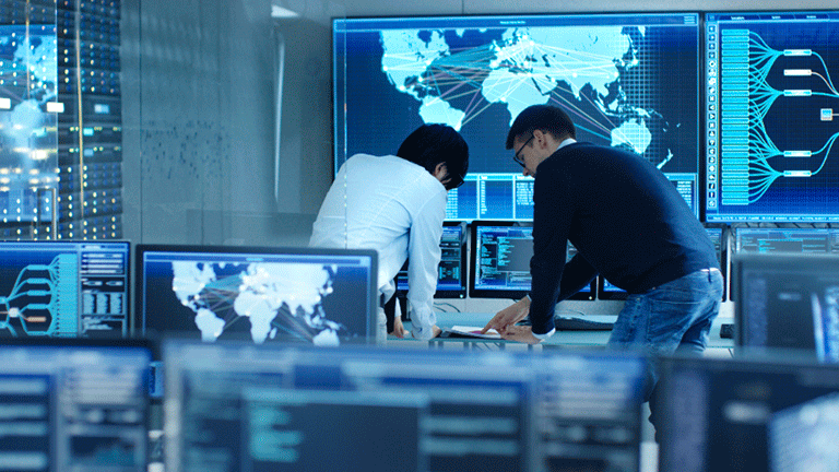 Two male coworkers collaborating in security command center with computer monitors in front and big screens in background