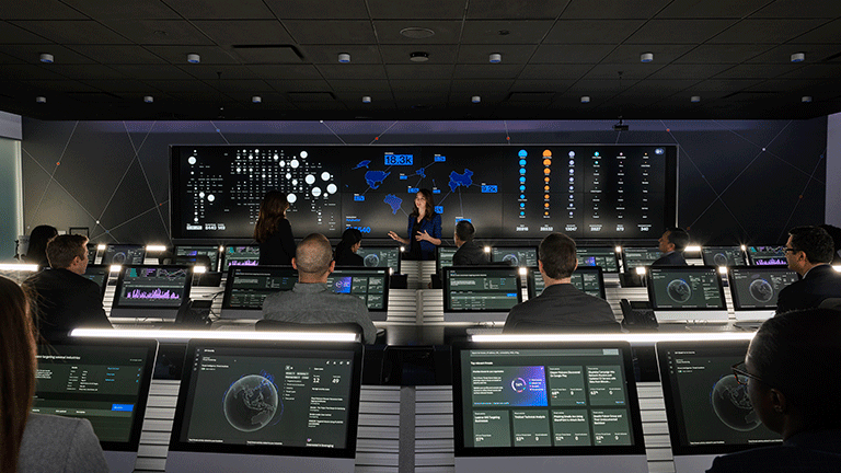 Woman presenting in front of coworkers in security command center with computer monitors and a large screen